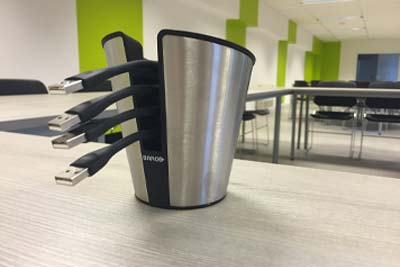 USB-Kabelhalter für Konferenzräume – Kabelmanagement Metallener Kabelhalter in Becherform mit mehreren herausragenden USB-Kabeln, auf einem Konferenztisch in einem modernen Meetingraum.